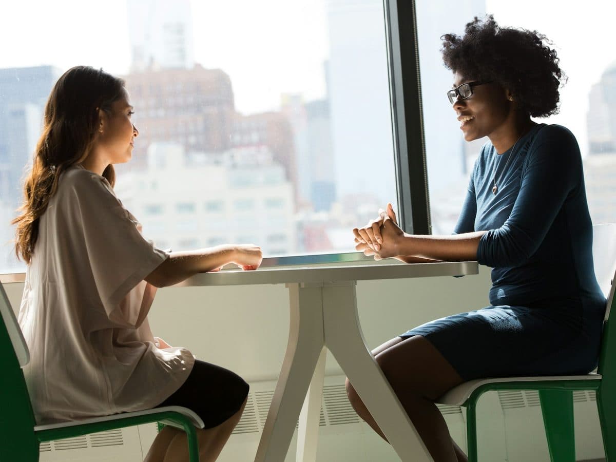 Two women talking across a small table by a window
