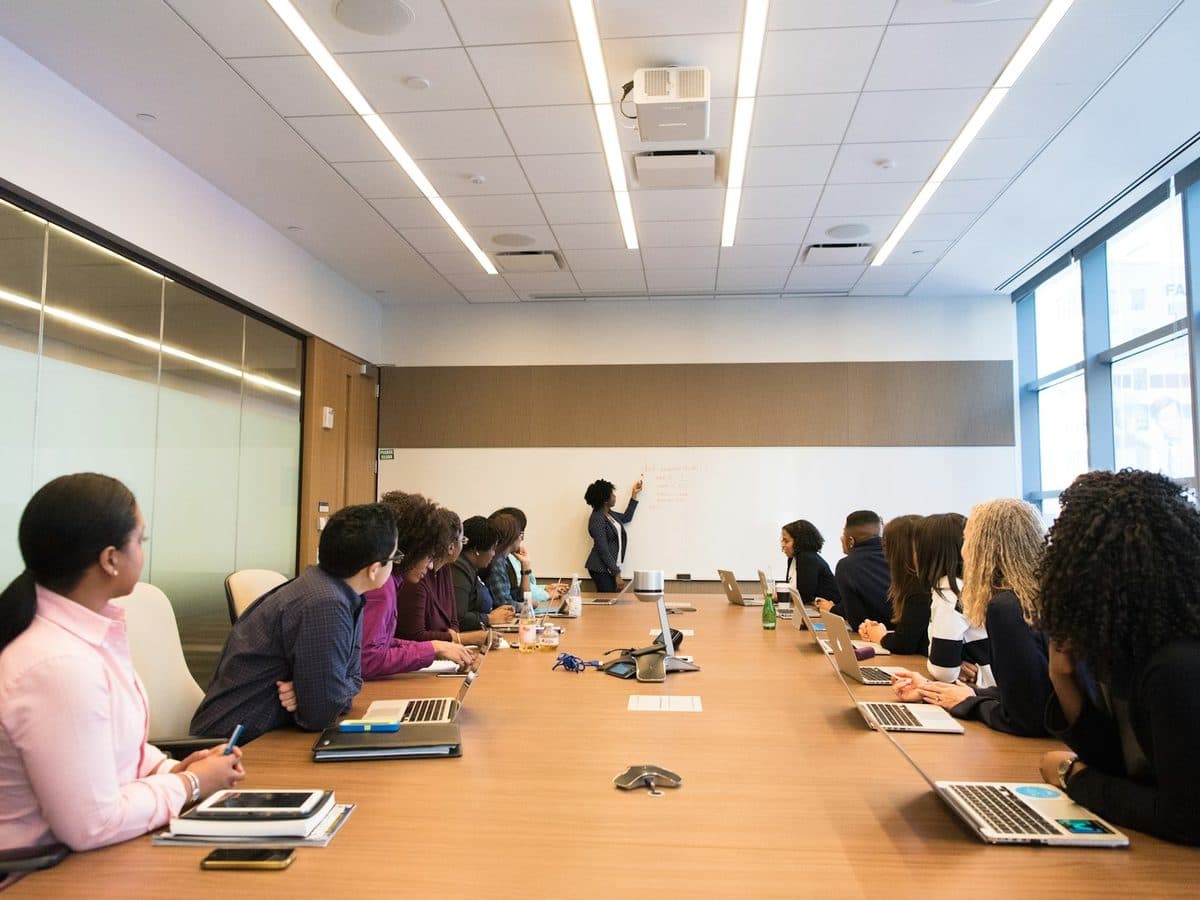 A trainer at a whiteboard with a room of practitioners around a long table