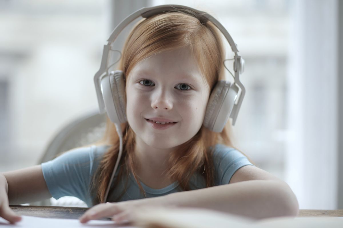 A smiling girl wearing white headphones, sitting at a table by a window